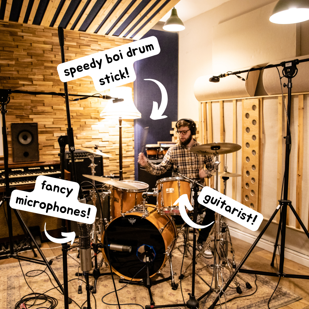 Luke playing drums in a recording studio. Text popouts on the image point to different picture features. #1 points to a blurry drumstick in Luke's hand and reads 'Speedy Boi Drum stick'. #2 points to Luke and reads 'guitarist!'. #3 points to 2 Aston Spirit microphones in XY position and reads 'Fancy Mics! 